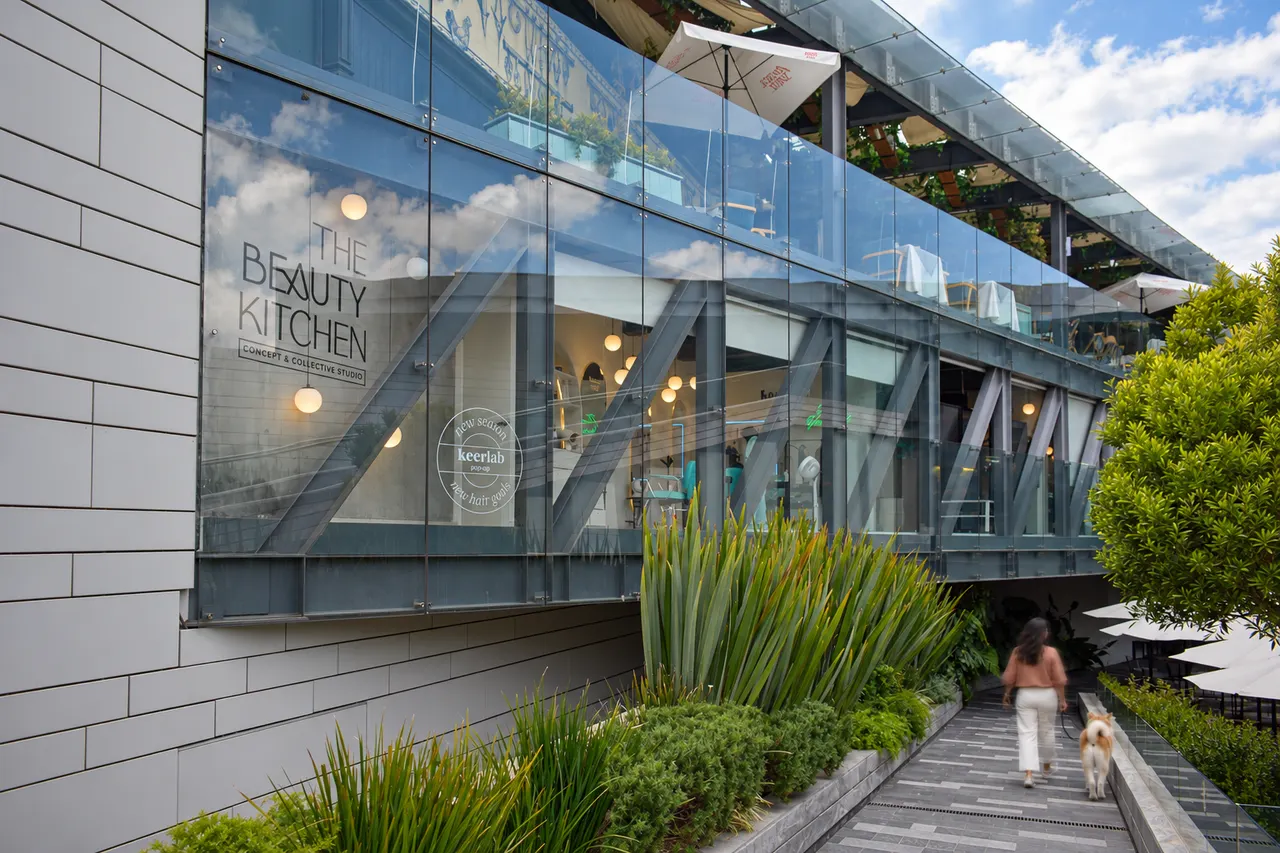 Fachada de cristal de The Beauty Kitchen y el pasillo con plantas por donde camina una mujer con su perro